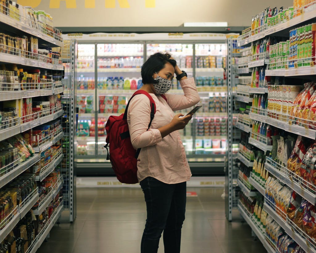 Woman stands in supermarket aisle looking at tinned options. In one hand she had her mobile phone and the other hand is pushing her hair away from her face as if she is concentrating.