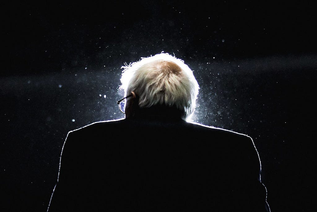 Senator Bernie Sanders’ behind a podium backlit by a bright light with particles in the air.