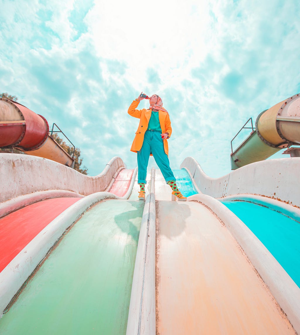 A woman standing on a pool slide that’s been out of order and drinking beverage.