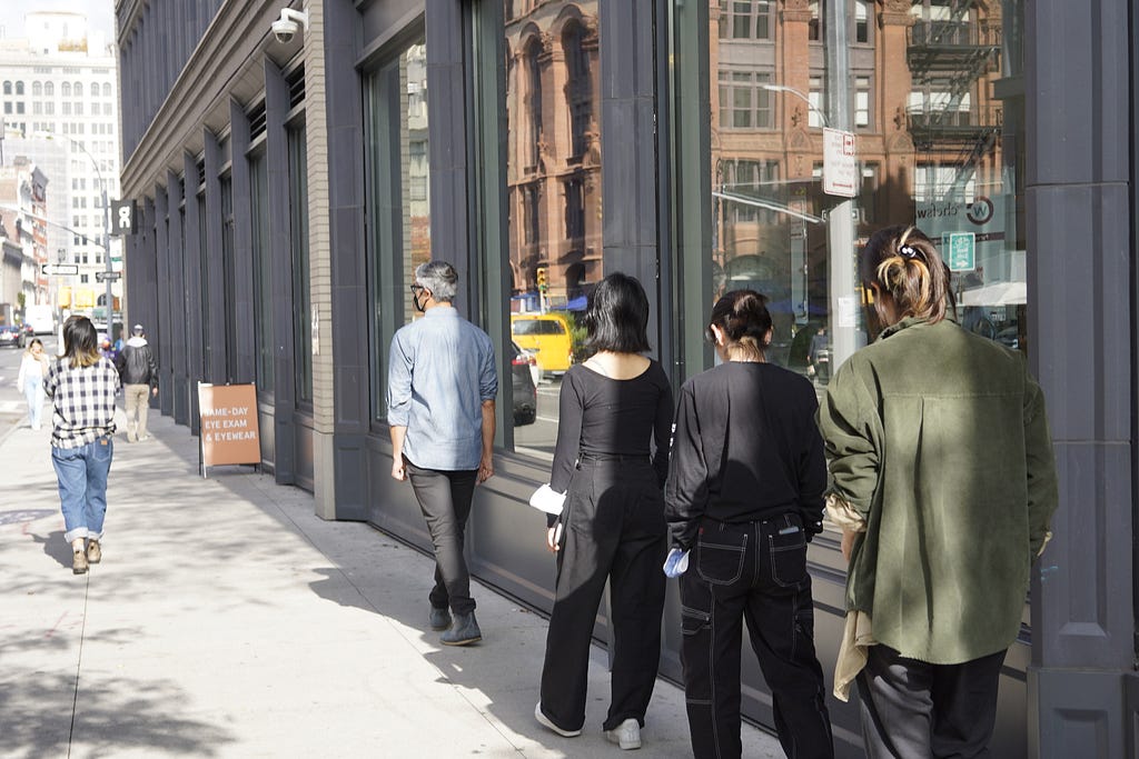 Participants follow Ann on a NYC sidewalk on a sunny day, walking in a single file in the shade alongside a shop’s windows.