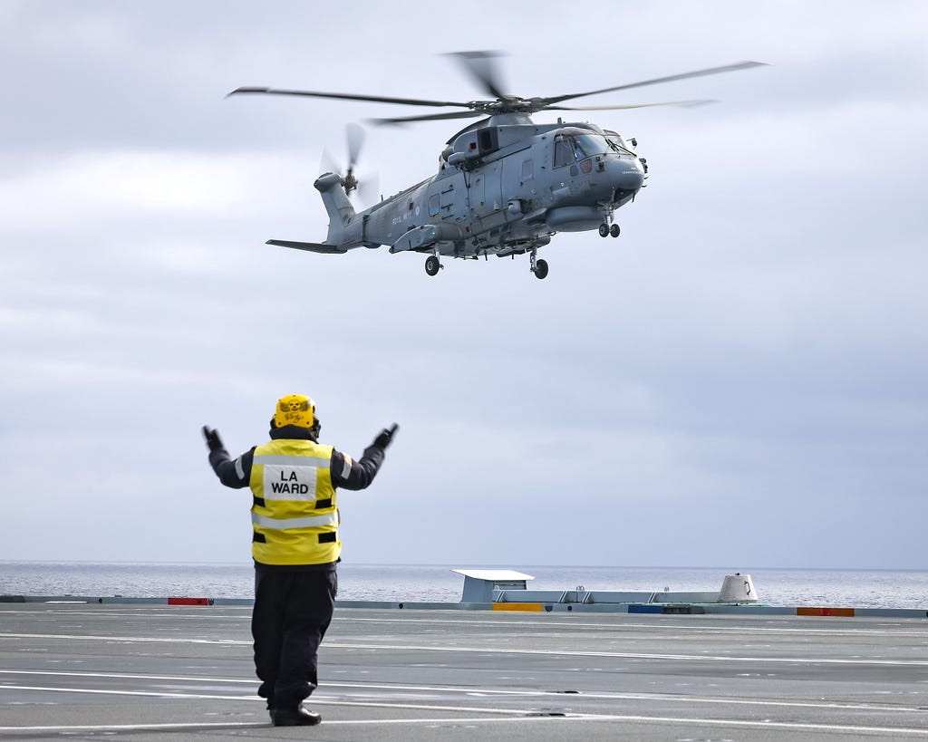A Merlin MK2 lands on HMS Prince of Wales with the help of a flight deck crew member