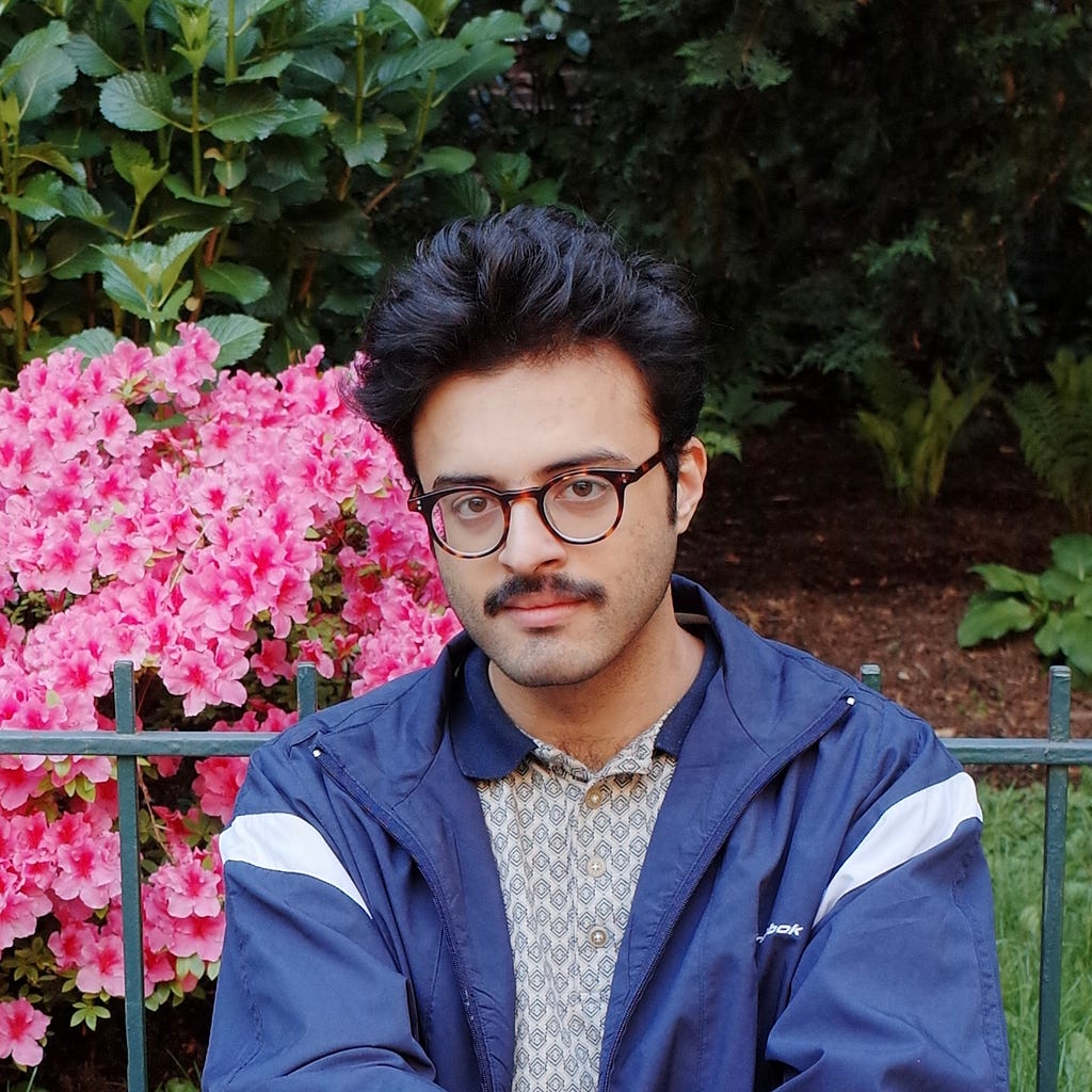 Amad seated against a green metal fence with pink flowers and green bushes in the background in Jackson Heights, Queens, wearing a patterned collared shirt and navy windbreaker.