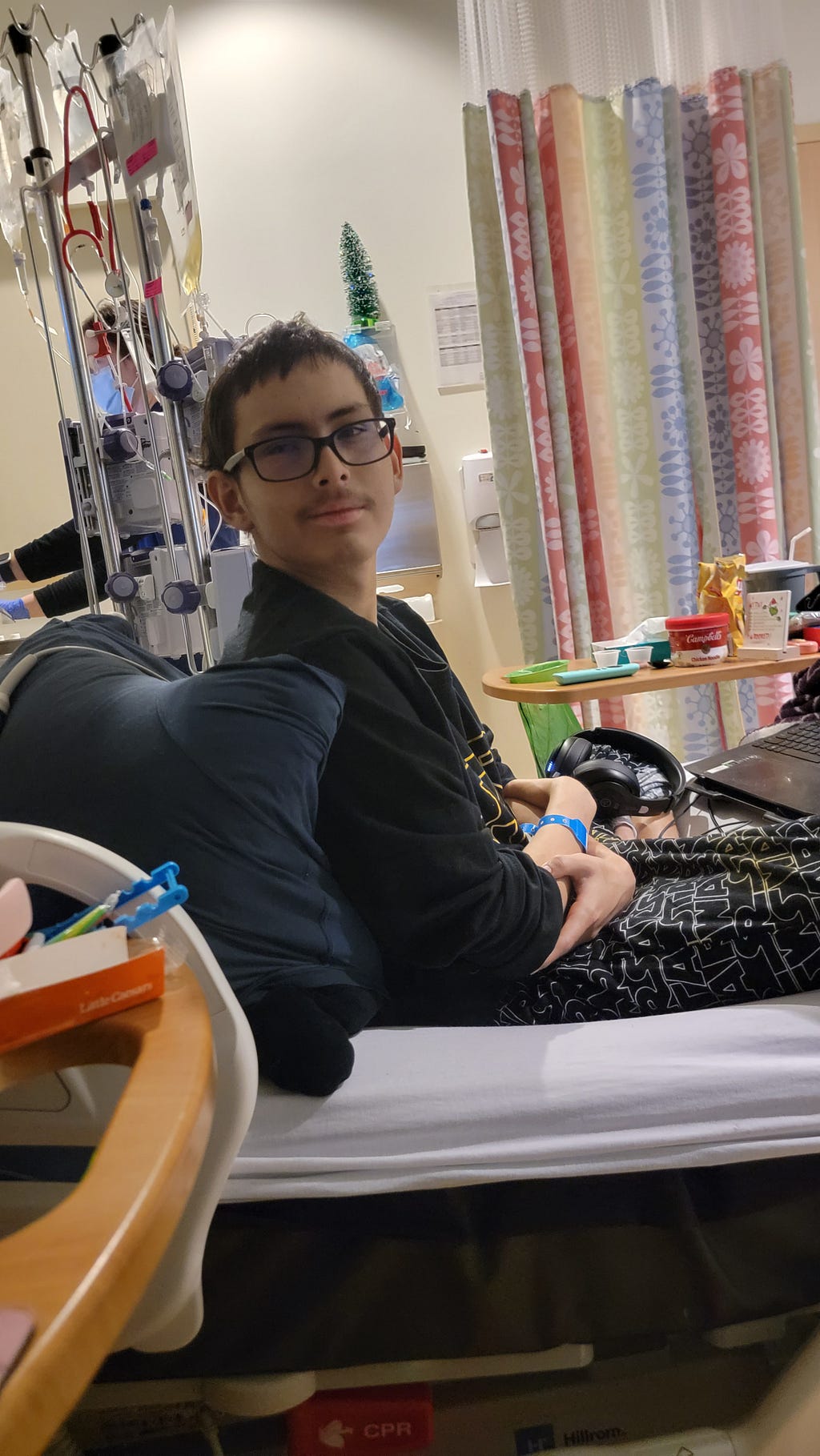 A person wearing glasses is sitting upright in a hospital bed, smiling. Medical equipment surrounds them. The room is softly lit and colorful curtains hang nearby.