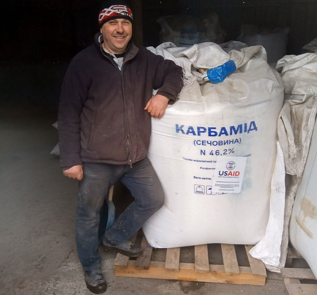 A man leans on a large bag that contains fertilizer, marked with the USAID logo.