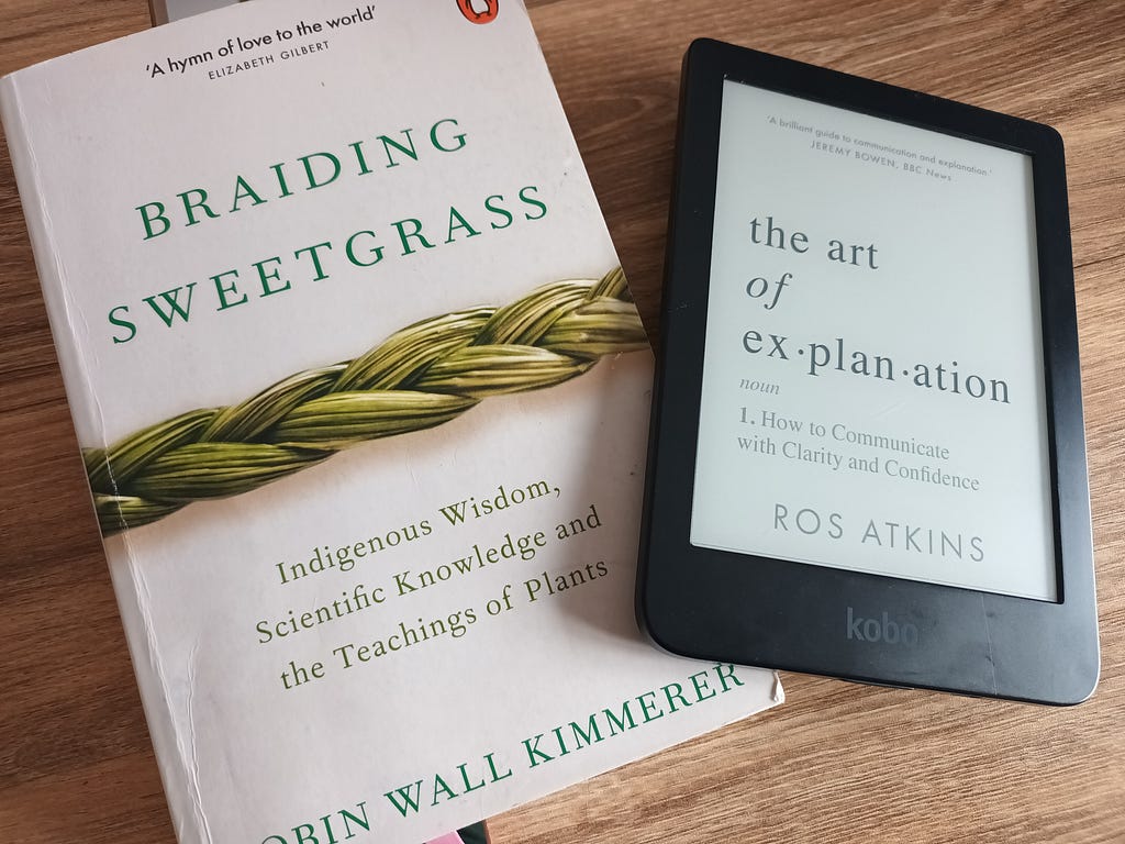 A paperback copy of Braiding Sweetgrass by Robin Wall Kimmerer and an e-reader showing the cover of the art of ex-plan.ation by Ros Atkins.