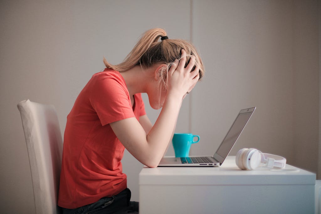 A blonde woman sitting in front of a desk, with her laptop open. She has her hands up in her head, covering her eyes, looking very confused and tired.