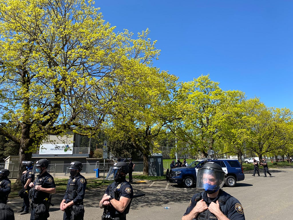 A line of police across the bottom of the photo. Behind them are a row of trees in Lents Park. A police SUV and more officers are visible in the background. Blue sky appears above the scene.
