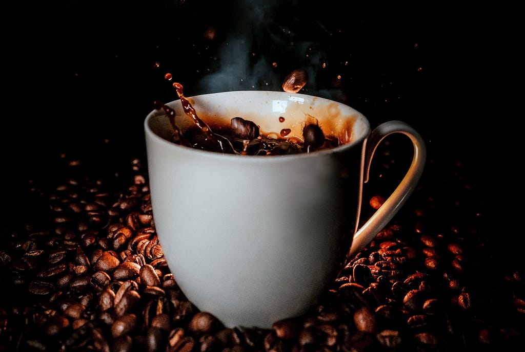 A white coffee cup filled with steaming coffee, sitting on a pile of coffee beans, with coffee beans splashing into the coffee.