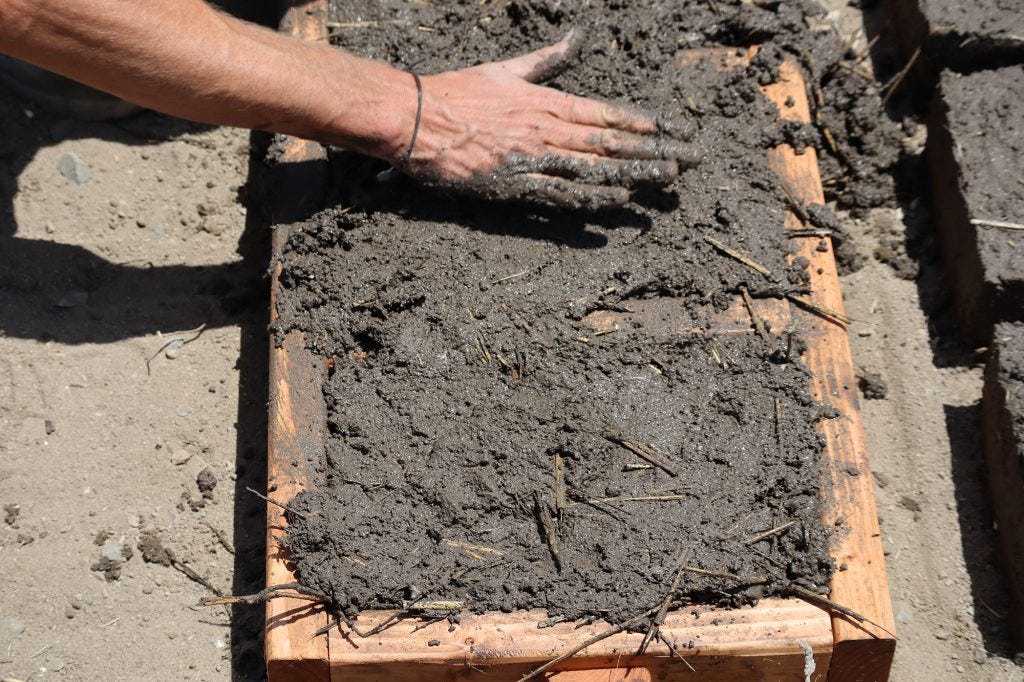A hand pats down the mixture of local soil, water, and straw into a wooden mold to create a cob brick. It will later be left to dry in the sun.