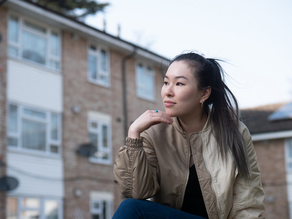 A young person is sitting outside in front of an apartment building. They are Asian and have light skin and long, dark hair tied into a ponytail. They are wearing a brown bomber jacket, and are looking off into the distance with their chin resting on their fingers.