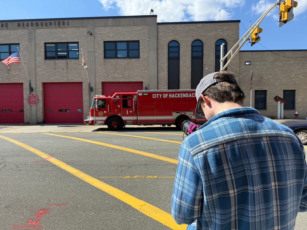 A man films outside of Hackensack Fire Headquarters with a firetruck rolling by.