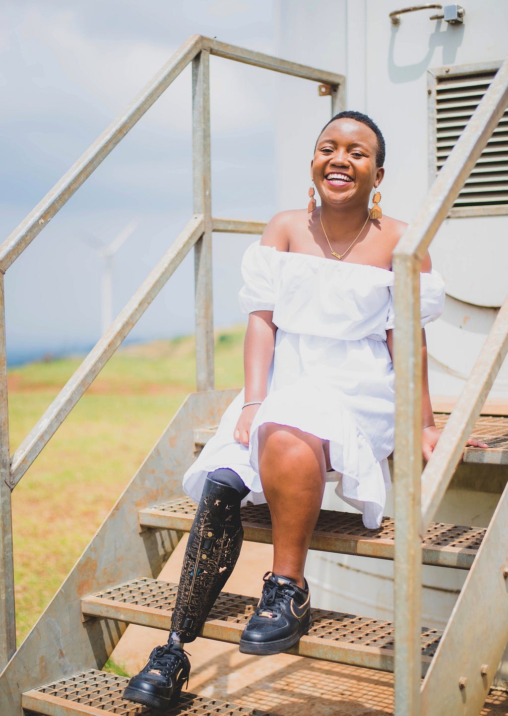 A picture of Elizabeth Mang’eni sitting on stairs, smiling. She is wearing a white dress and golden earrings.