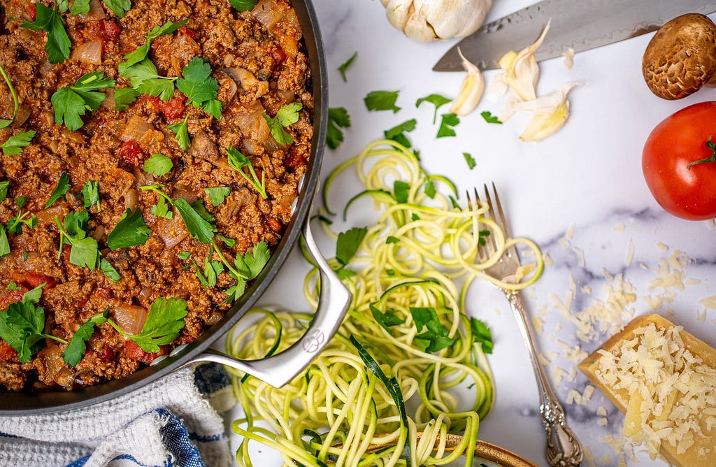 A large pan of cooked ground beef surrounded by garlic, zucchini, tomatoes, and parmesan cheese.