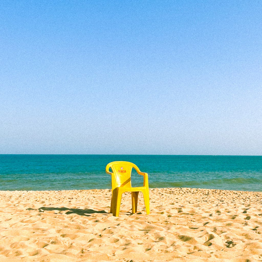 Uma cadeira amarela de plástico na praia, com o mar no fundo dividindo a paisagem com o céu. Foto: Carol Poleze.