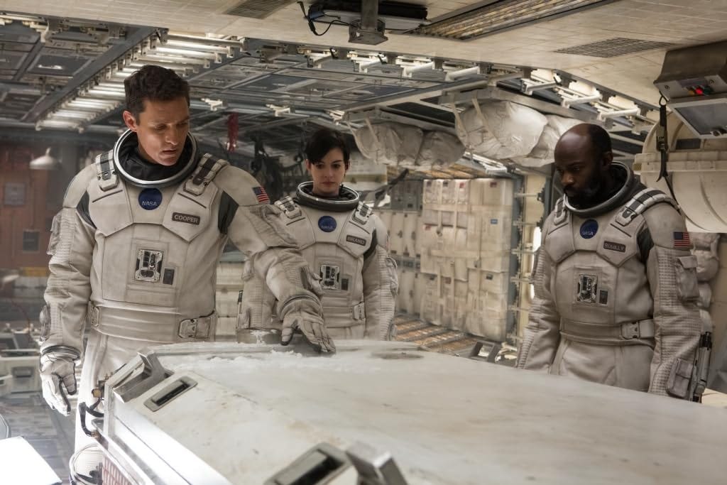 IMDB still from Christopher Nolan’s “Interstellar” showing a white male, white female, and black male in space suits examining a high-tech sleeper container on a futuristic space ship. https://www.imdb.com/title/tt0816692/mediaviewer/rm2319369216