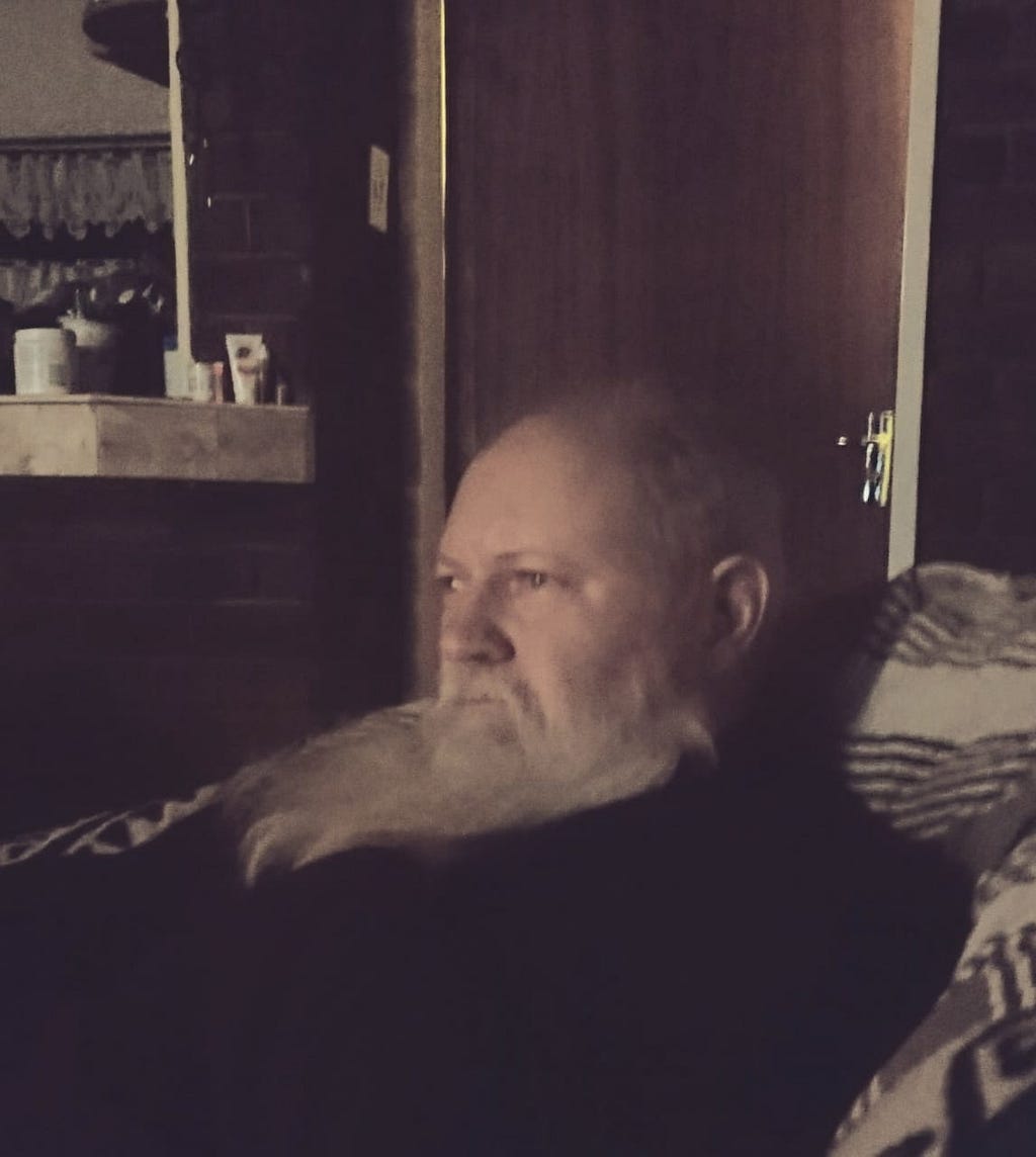 A Caucasian male with a long grey beard and bald head sitting on a chair in a living room and staring into the distance pensively