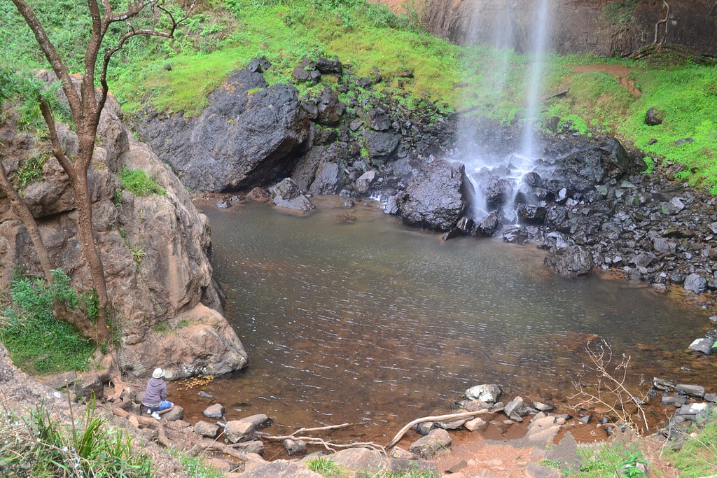 A person sitting at a rock looking at the edge of a rock pool at the base of a waterfall