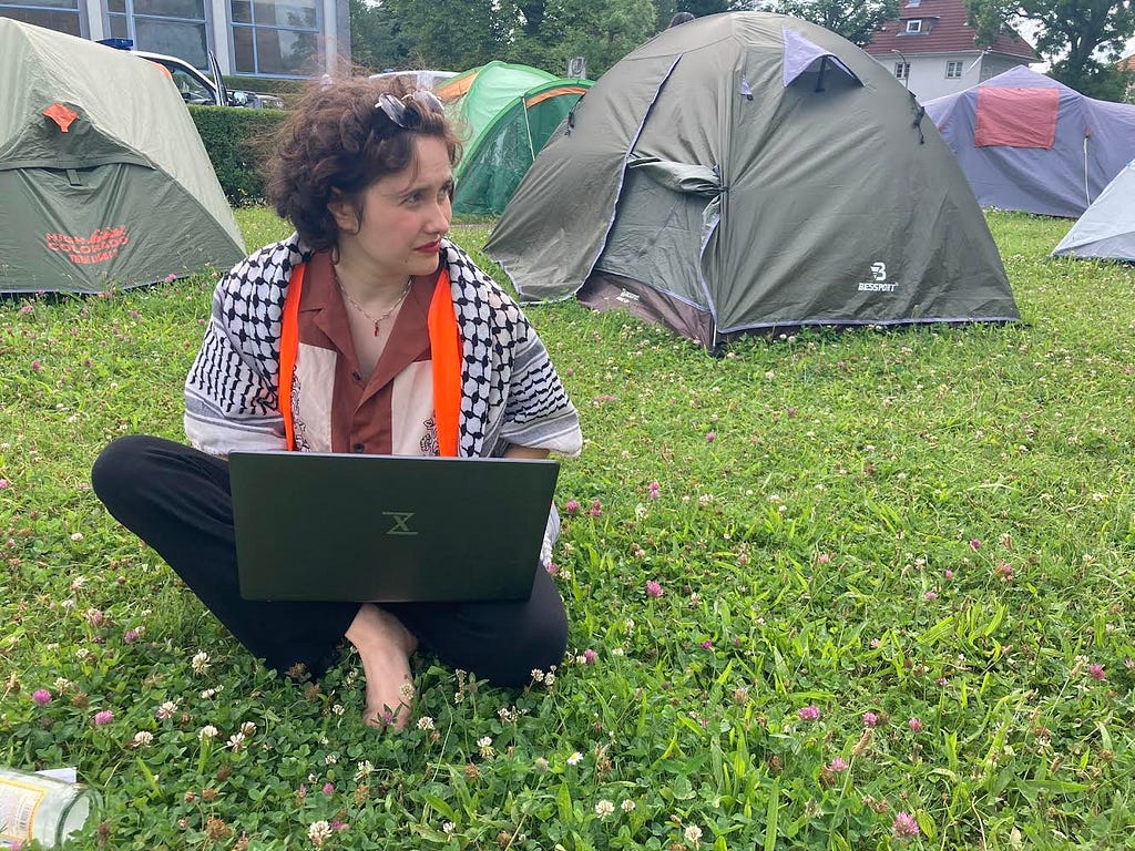 A young student draped in a keffiyeh sits on the grass outside a set of tents