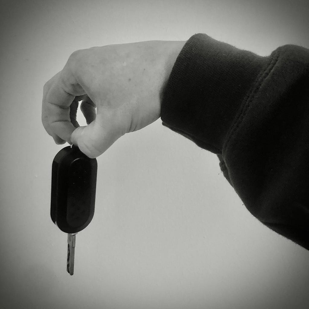 A black and white photo of an arm outstretched, dangling  a car key in their fingers.