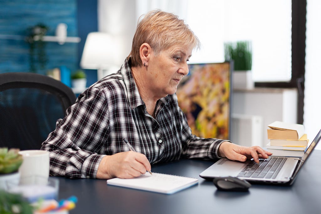 Old woman navigating on a laptop and writing on a notebook.