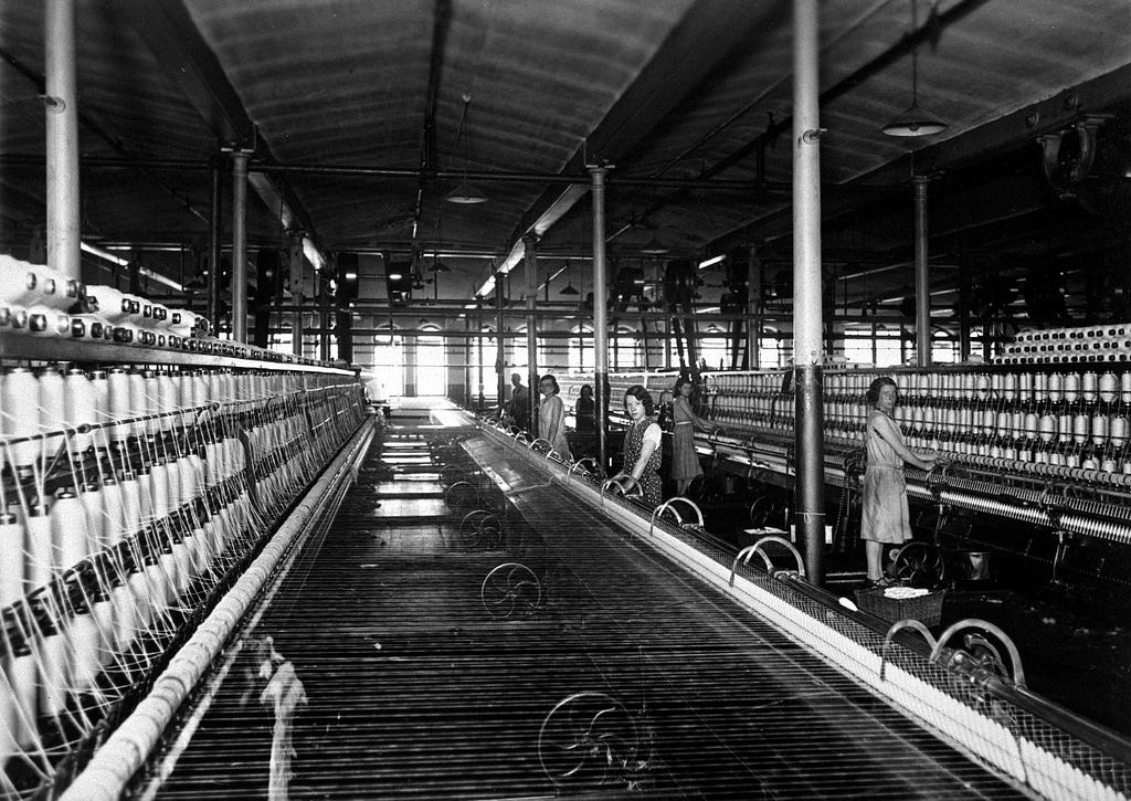 Large spinning room where many young women and children are seen working. The child on the far right is so small she has to stand on a basket in order to reach.