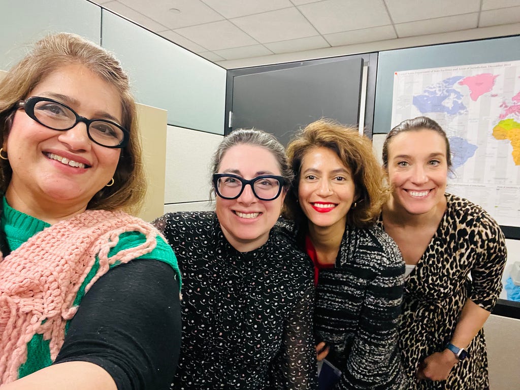 Four smiling women squeeze into an office cubicle.