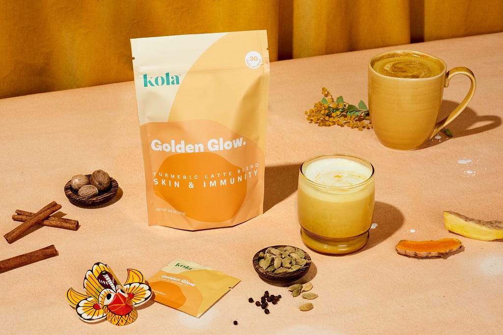 A Kola Goodies Golden Glow pouch with spices, sri lankan masks, mugs of turmeric latte in a in a yellow/golden background