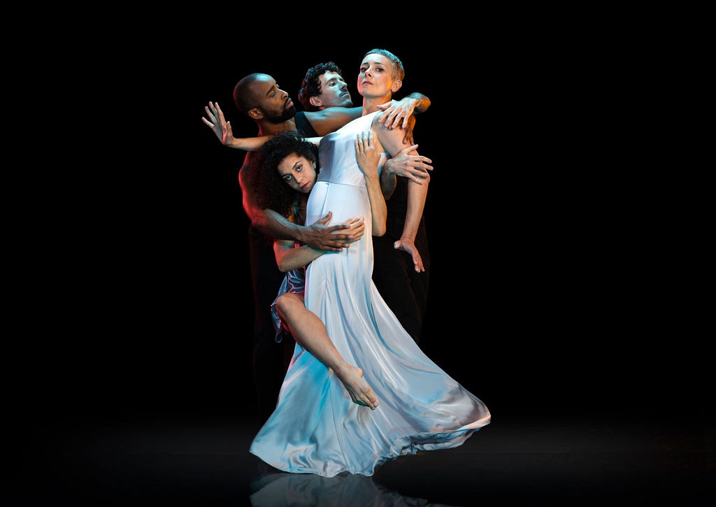 A group of dancers perform on a black background. Three dancers hold on to another dancer, who wears a long white dress.