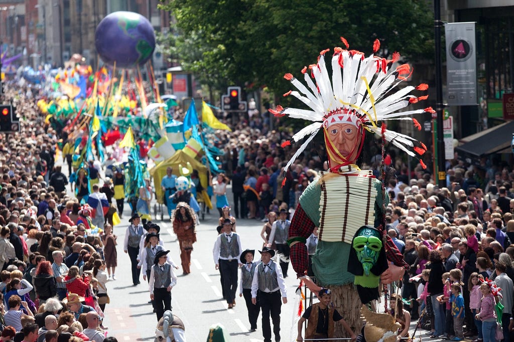 A wide shot of the Manchester Day Parade with musicians and large puppets parading down the street, framed on either side by crowds of onlookers.