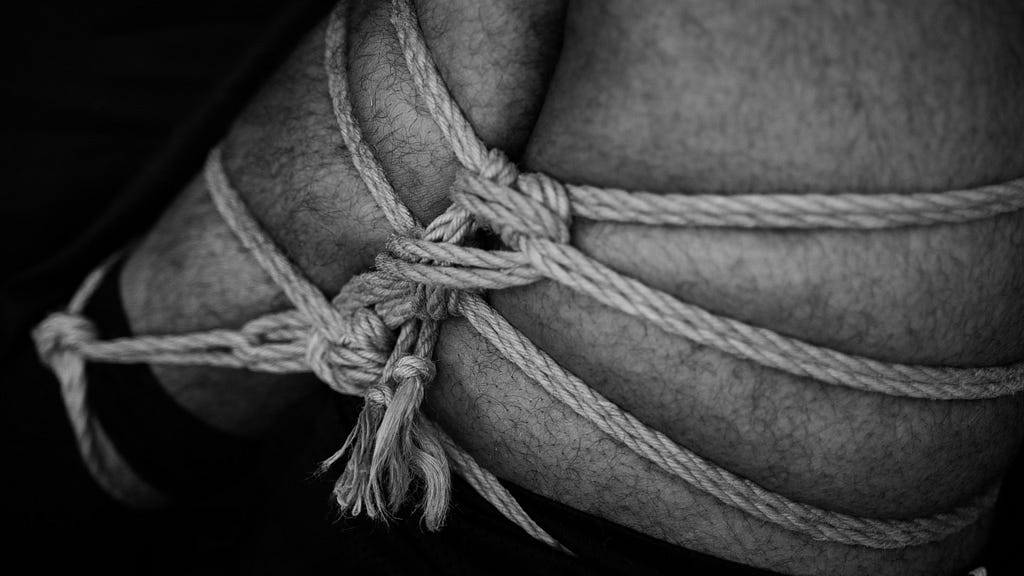 A man’s leg is tied ornately with rope and elaborate knots. Photo is black and white.