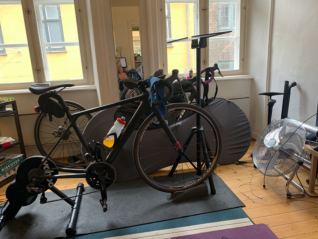 A road bicycle attached to a ‘wheel-off’ smart trainer, with a music stand for ipad, a large floor fan, on top of a yoga mat to catch sweat