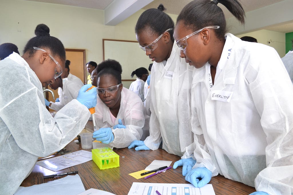 One of four women wearing white lab coats, goggles, and light blue disposable gloves demonstrates a laboratory technique to three of her fellow researchers.