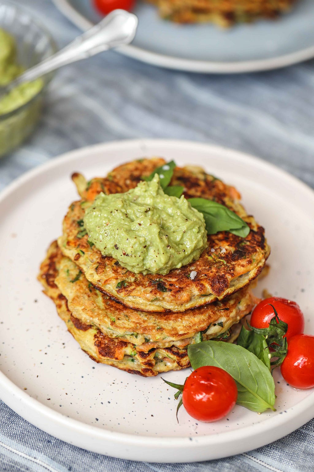 Three stacked zucchini fritters, topped with avocado sauce, and garnished with basil and cherry tomatoes, by FIT & NU