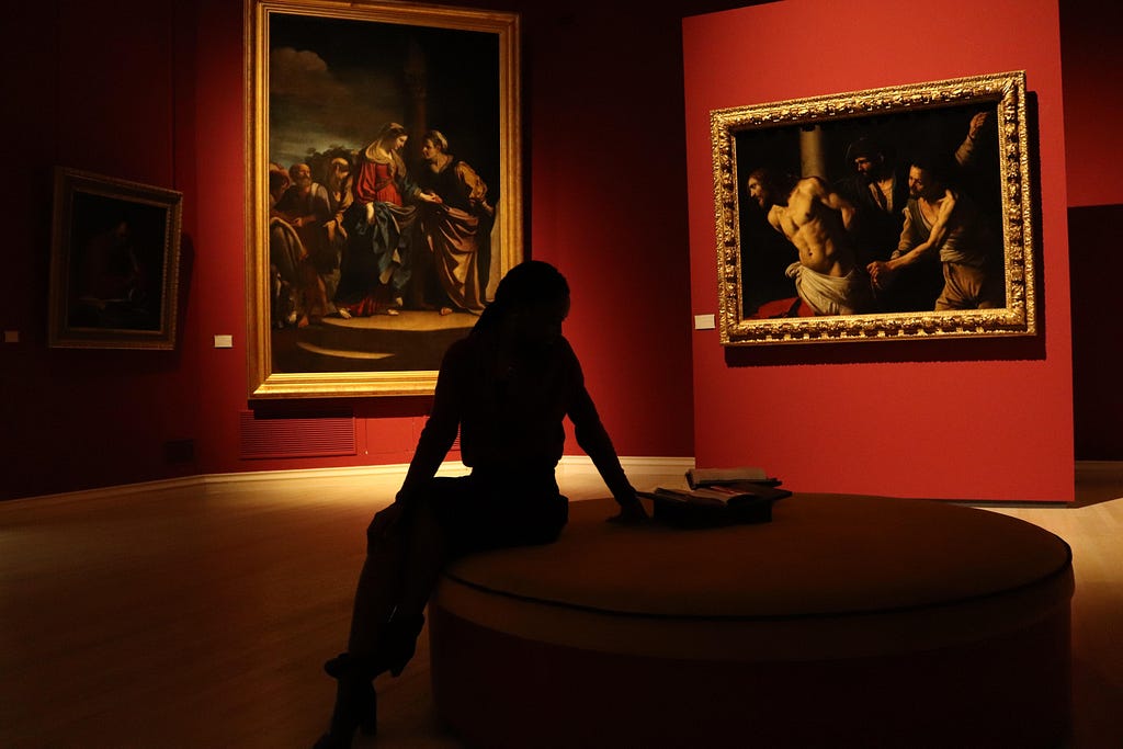 A black silhouette of a person sitting in front of two paintings which are illuminated by artificial light in a museum setting.