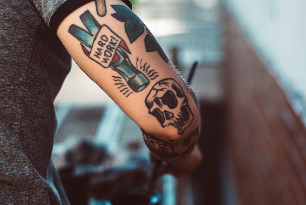 Picture of arm with tattoos, one says “Hard Work” on it.