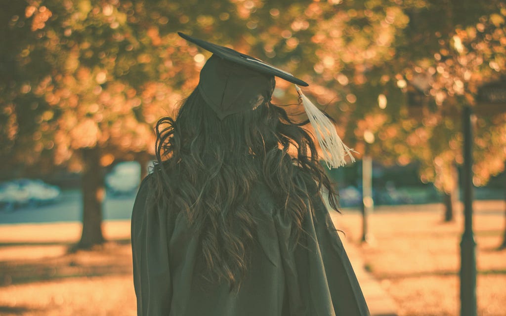 A woman wears a black graduation cap and gown with a gold tassel. She is walking away from the camera. In the background are several trees with fall leaves.