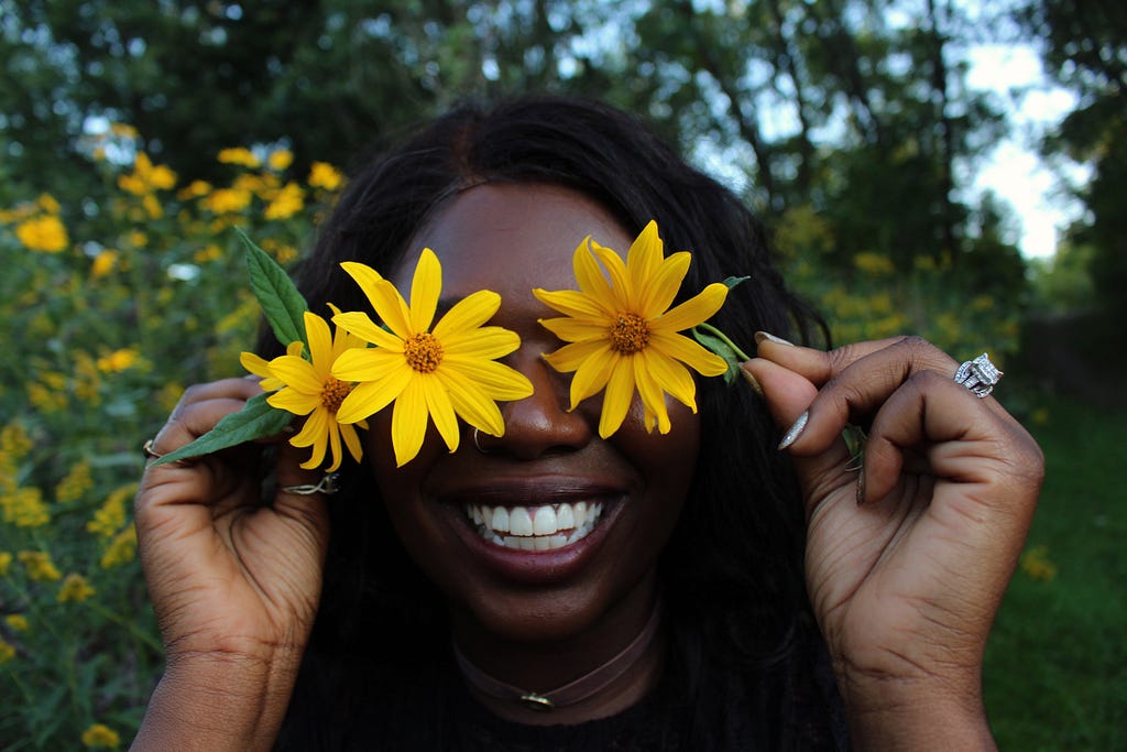 An African American woman holding up flowers in front of her eyes in the park.