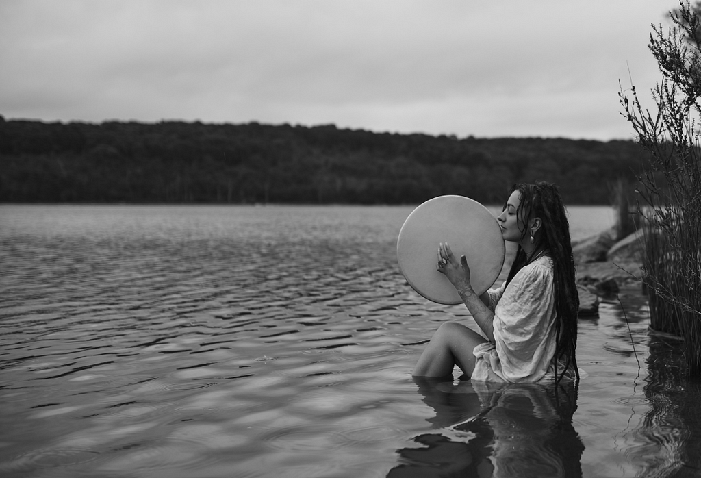 A woman sits in the shallow waters of a lake, holding a large drum up to her chin with both hands.