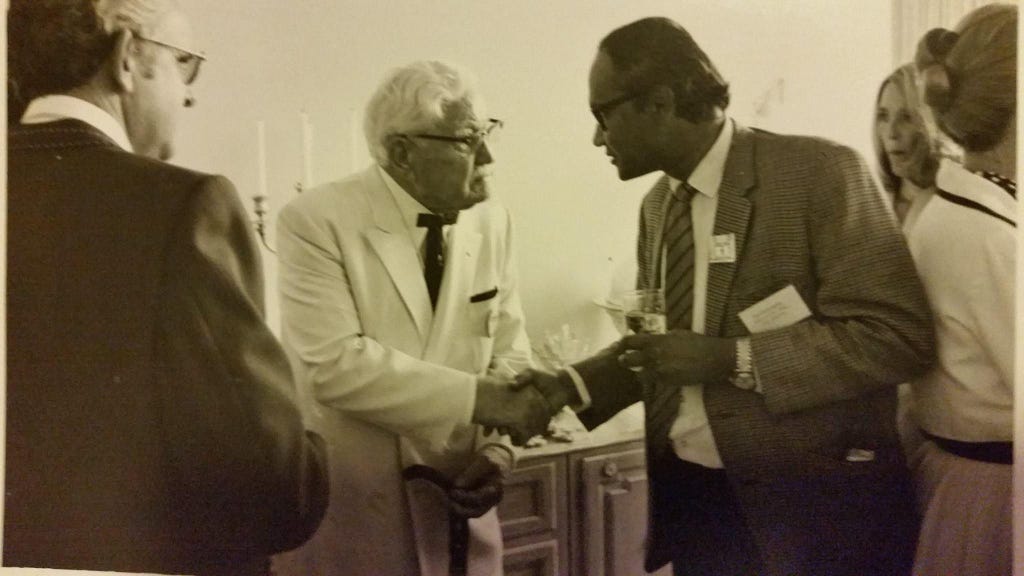 Colonel Sanders is depicted in an old black-and-white picture standing on the left in his traditional attire of a white suit and a black bowtie, holding a cane. My father stands on the right in a business suit, holding a drink, shaking hand for the photograph.