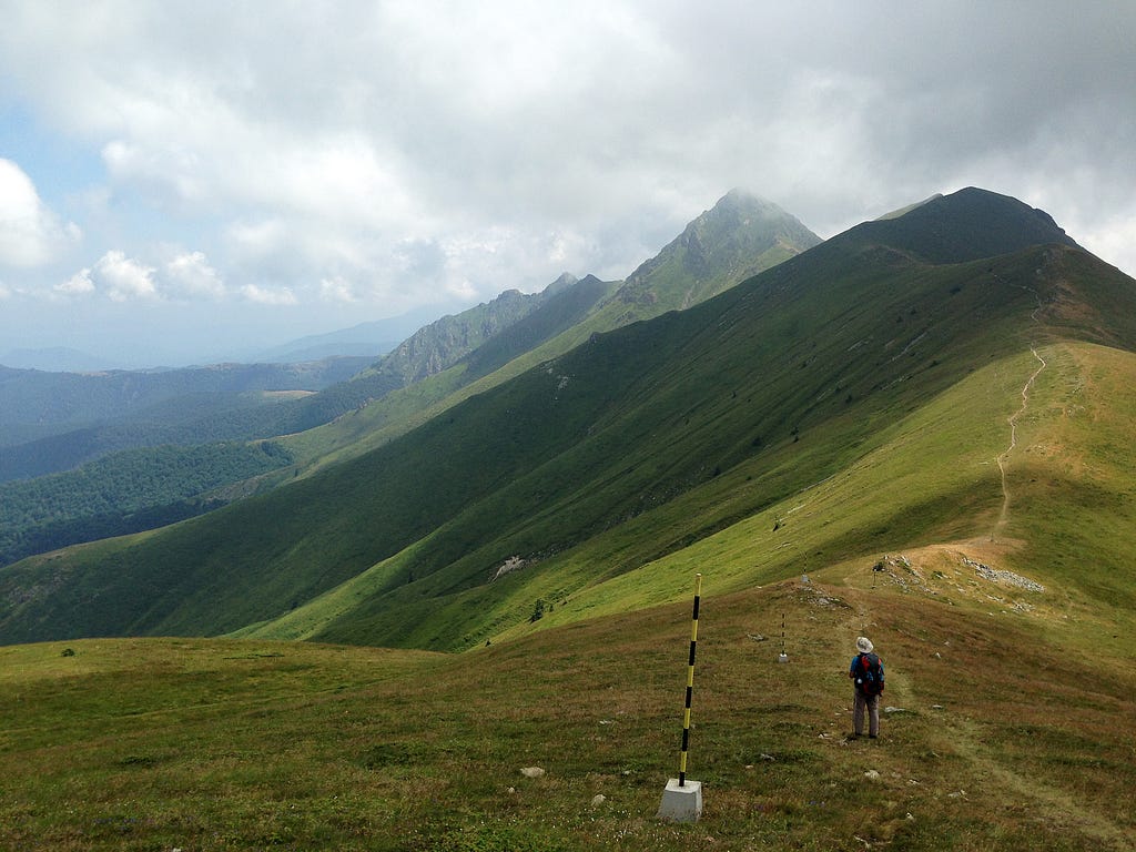 A green mountain range, in the foreground is a hiker. In the distance a path goes on the ridge, peaks are in clouds.