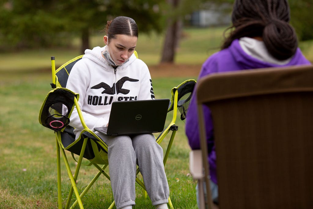 Two female high school students sit in lawn chairs outside, working on laptops.