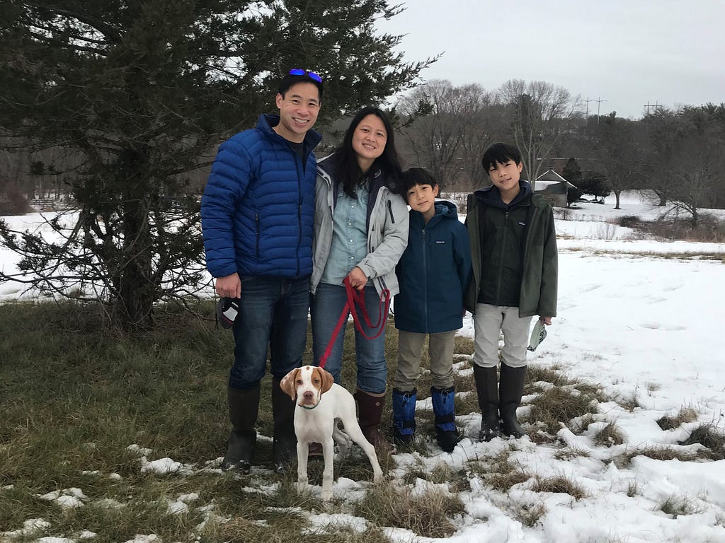 A man, woman, two boys, and dog stand in the snow with a tree behind.