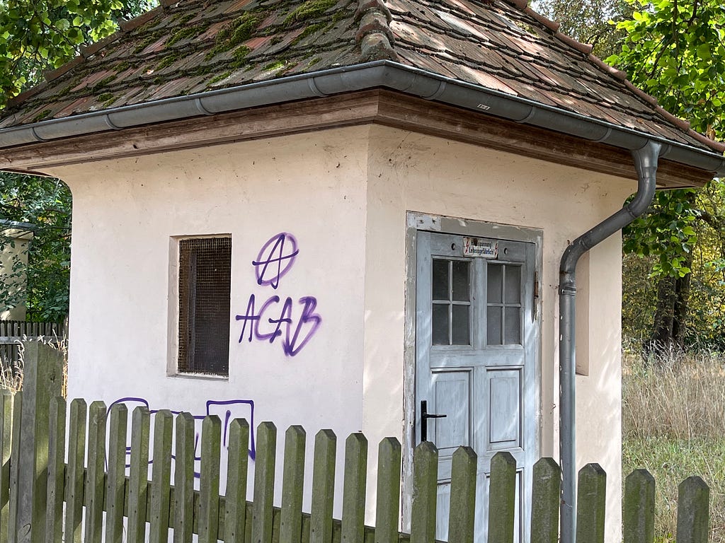 Photograph of a small, light pink structure with one door and window behind a wooden fence. On the side of the building, ACAB and the anarchy symbol have been spray-painted in dark font.