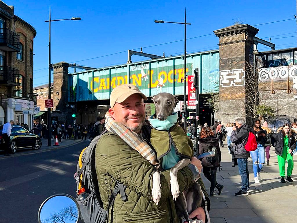 A man in a green jacket is holding a whippet, also in a green jacket, in front of Camden Lock in London