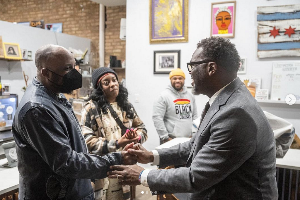 Brandon Johnson (in a gray suit) shakes my hand at a coffeeshop community event. I (a black Black man) am in a KN95 mask and a blue fake-leather jacket. In the background, two other Black community members look on with admiration on their faces.