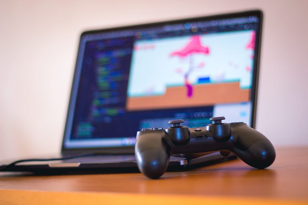 The image depicts a video game controller in the foreground, connected to a laptop in the background. On the laptop screen, we can see a video game