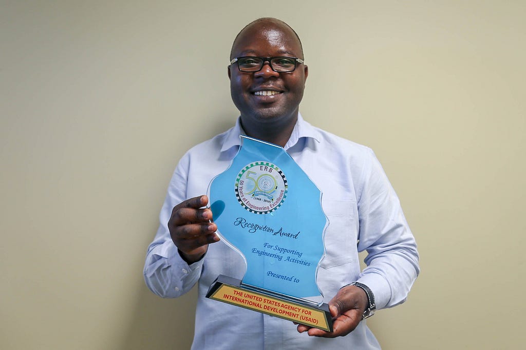 A man holds an award shaped like a splash of water that reads: Recognition Award For Supporting Engineering Activities Presented To USAID.