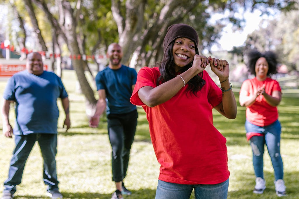 A park. Four people celebrating. A woman dancing in the foreground, she’s wearing a red shirt and jeans. Behind her another woman is wearing the same outfit. The two men that are also behind her are blue shirts and jeans.