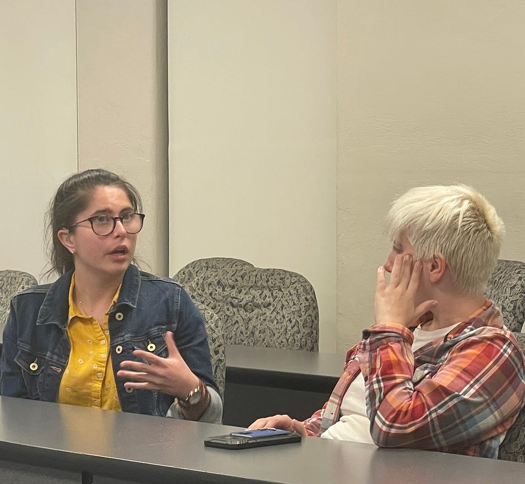 Two students talking in a classroom while sitting in rolling chairs.