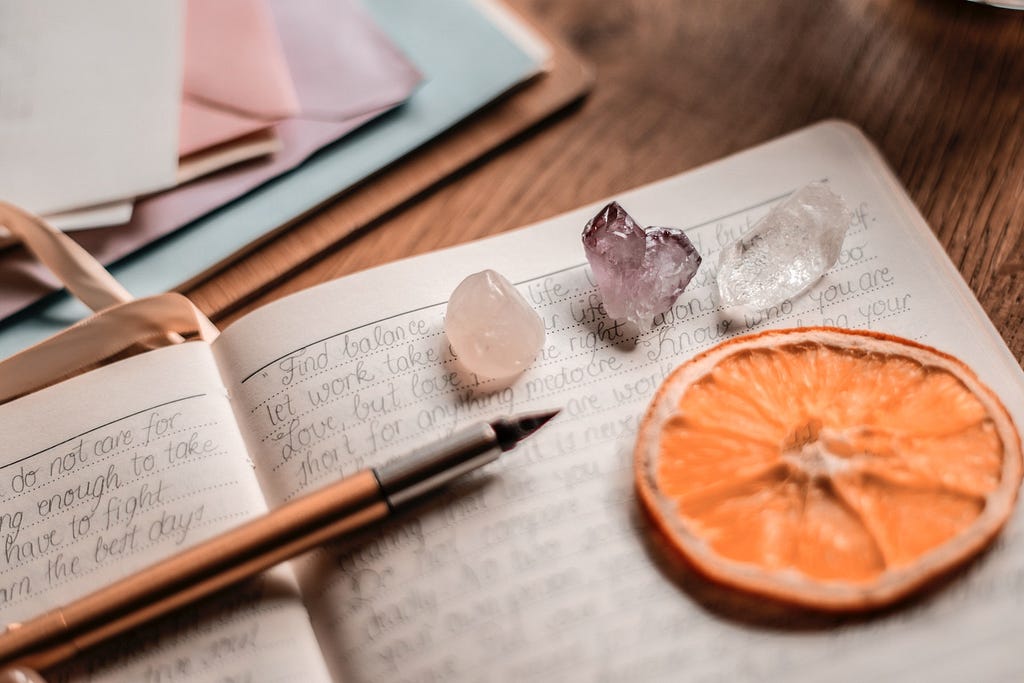 A a desk with a close-up of a journal, writings on pages, a pen lays across it. A dried orange slice and gemstones lays on one pages. Envelopes in pink, blue and purple above it.